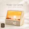 A wooden sunrise alarm clock with a built-in diorama showing a sunset over the ocean, placed on a light beige surface. The clock has a textured gray front panel with a digital LED display showing 7:00.
